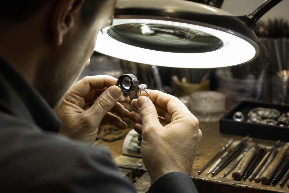 A jeweler's hands examining a custom-designed ring under bright magnification lamp, closely inspecting gemstone placement and metal finish with specialized loupe, showing meticulous attention to detail