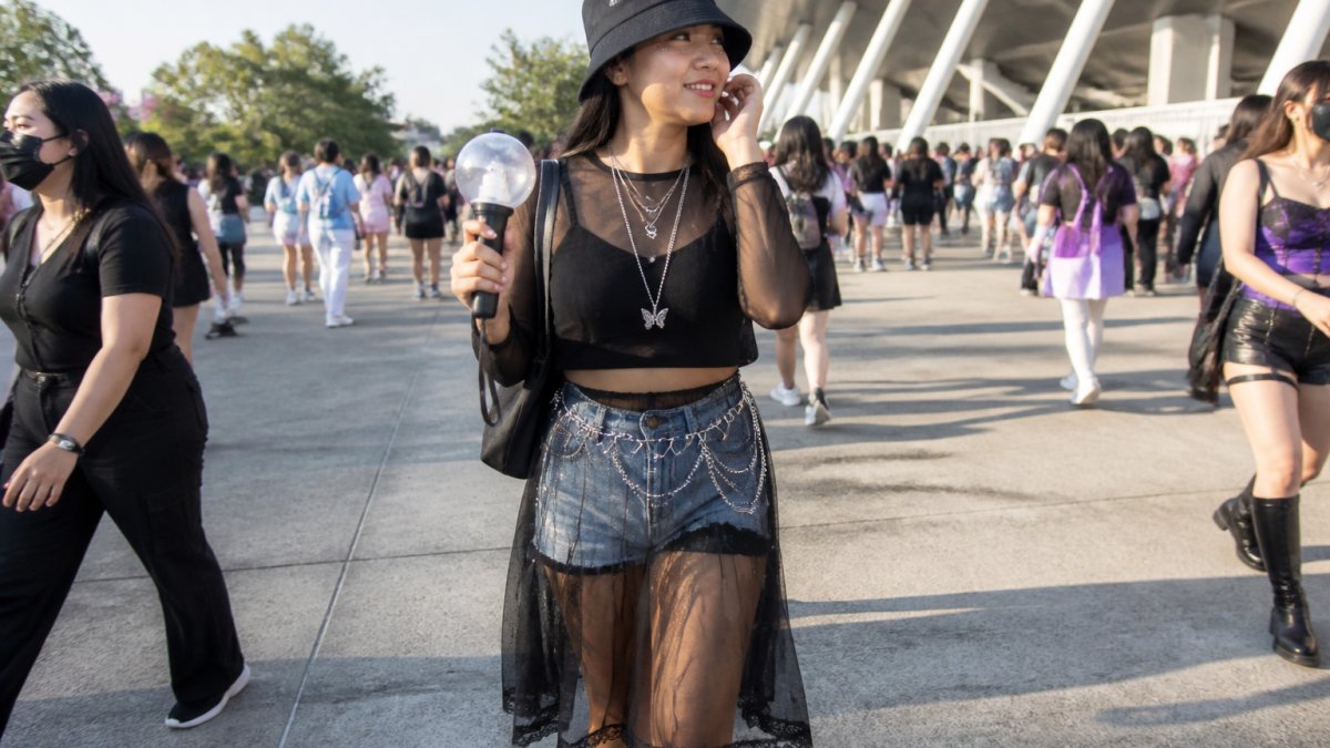A young K-pop fan in a sheer mesh skirt layered over denim shorts, wearing layered necklaces and a bucket hat, standing in a bright outdoor stadium line with an ARMY Bomb light stick in hand
