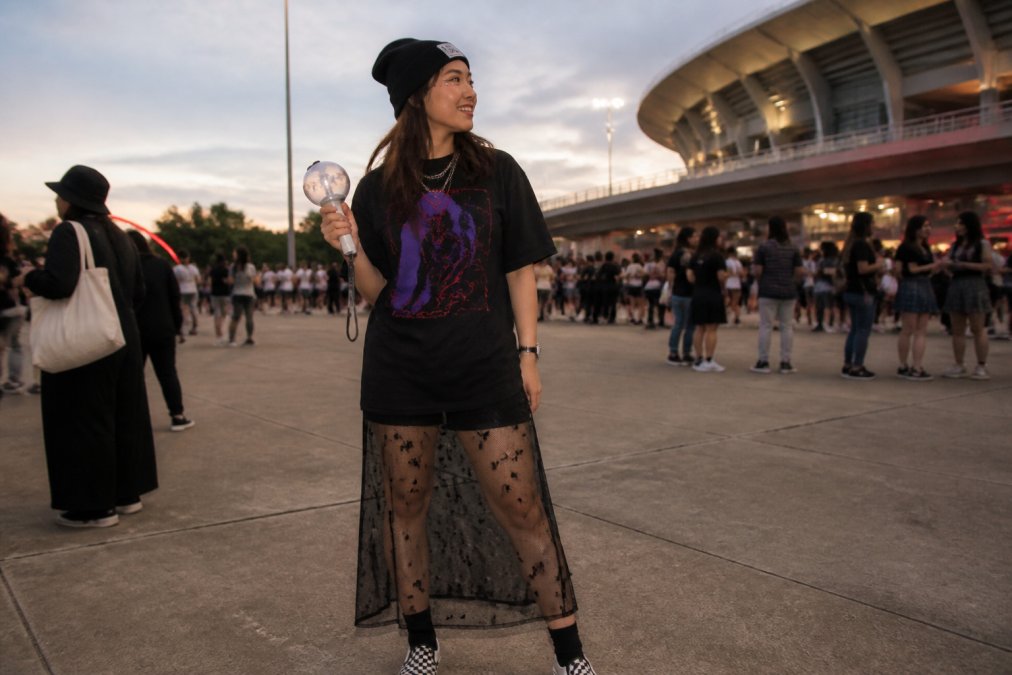 K-pop fan wearing custom graphic tee and sheer mesh skirt at outdoor concert venue, styled with beanie, slip-on shoes, and army bomb lightstick, vibrant purple and red graphics visible, natural evening lighting