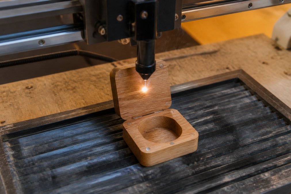 Close-up of a laser engraving machine precisely etching a wooden ring box on a workbench, soft warm workshop lighting, fine wood grain visible