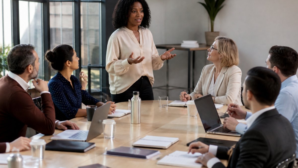 Professional facilitator leading a group of managers in an interactive workshop, seated around a large table with notepads and laptops, engaged in active discussion and collaborative problem-solving with natural office lighting