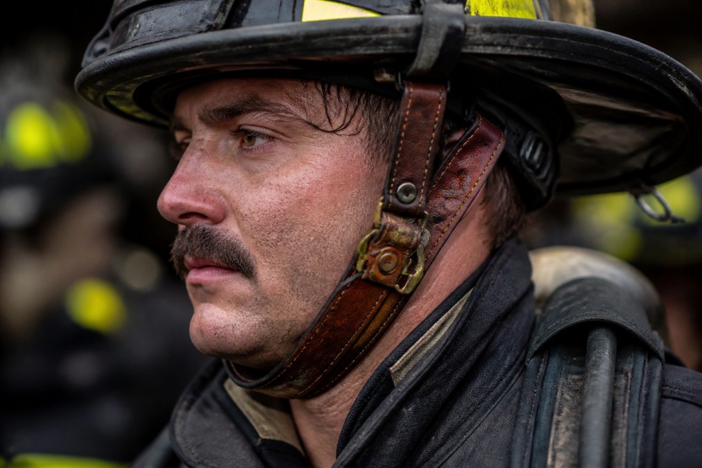 Close-up of a firefighter wearing custom leather helmet chin strap and front holder during active duty, showing rich patina and wear patterns on the leather surface under station lighting