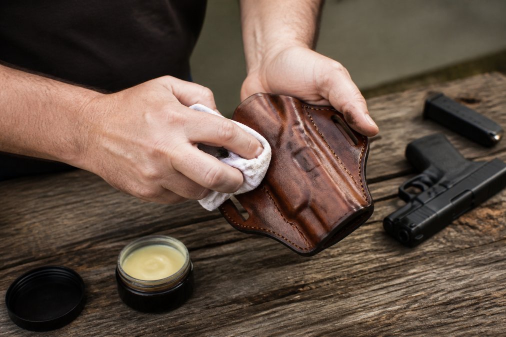 Hands cleaning and conditioning a leather holster with cloth and leather care product, showing maintenance technique with natural lighting on wooden surface