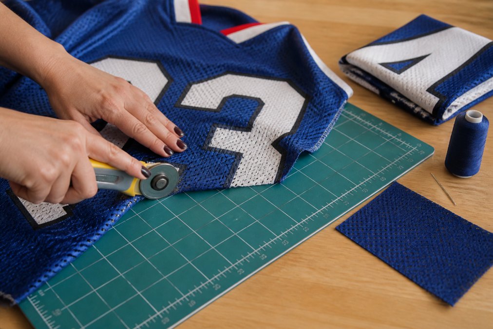 Hands carefully cutting vintage sports jersey fabric with rotary cutter on a self-healing mat, with marked measurements visible, thread and needle positioned nearby on a clean work surface