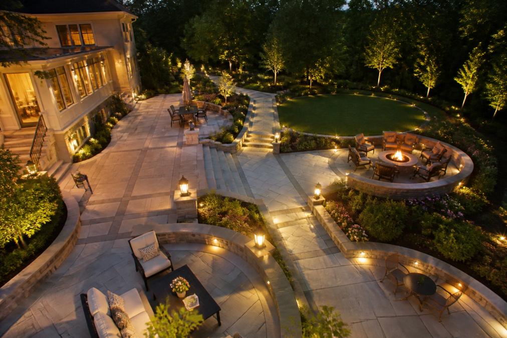 Wide aerial view of a luxury residential property featuring expansive custom stone patio with geometric pattern, multiple levels connected by masonry pathways, integrated landscape lighting casting warm shadows on stone surfaces, manicured gardens and trees framing the hardscaping