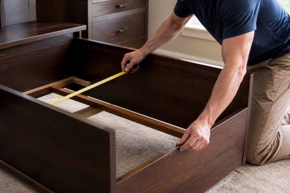 Close-up of someone carefully measuring interior bed frame dimensions with a steel tape measure, checking corners and frame depth with precision, in natural daylight