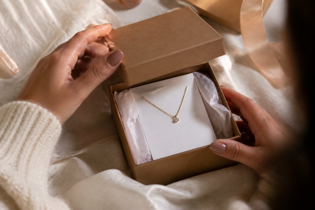 Hands carefully lifting a jewelry box lid to reveal a delicate necklace with a handwritten custom message card placed prominently on top, surrounded by soft tissue paper, warm natural lighting