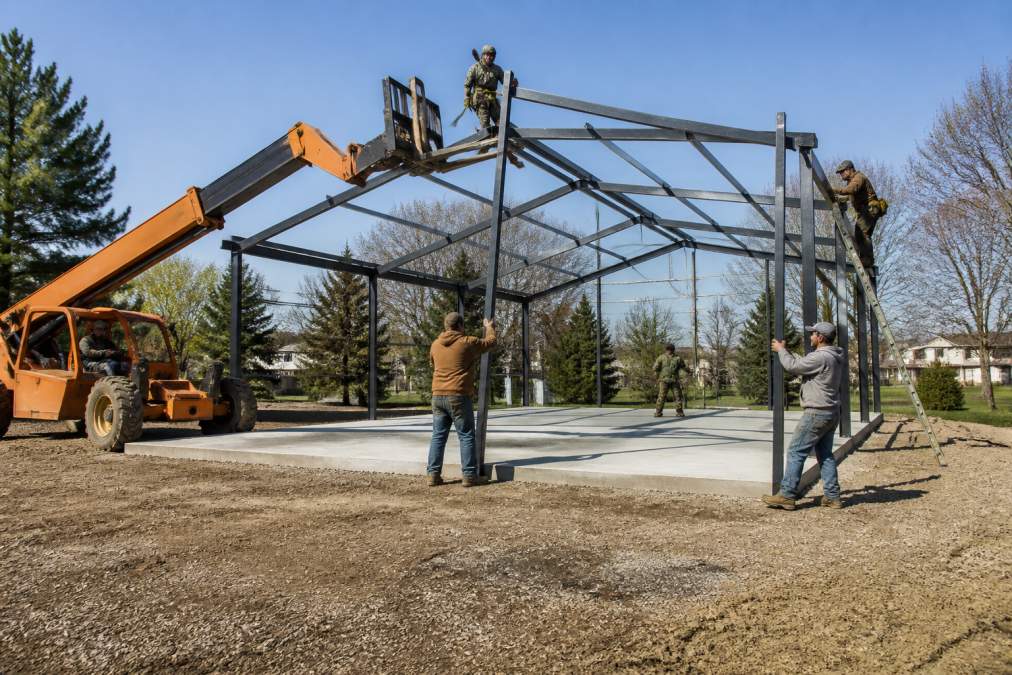 Installation crew securing a custom metal barn on a residential concrete foundation using heavy equipment