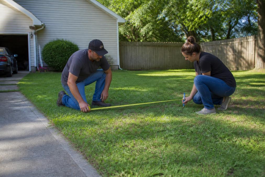 Homeowner measuring a residential lot with a tape measure, marking dimensions on property near driveway, natural daylight