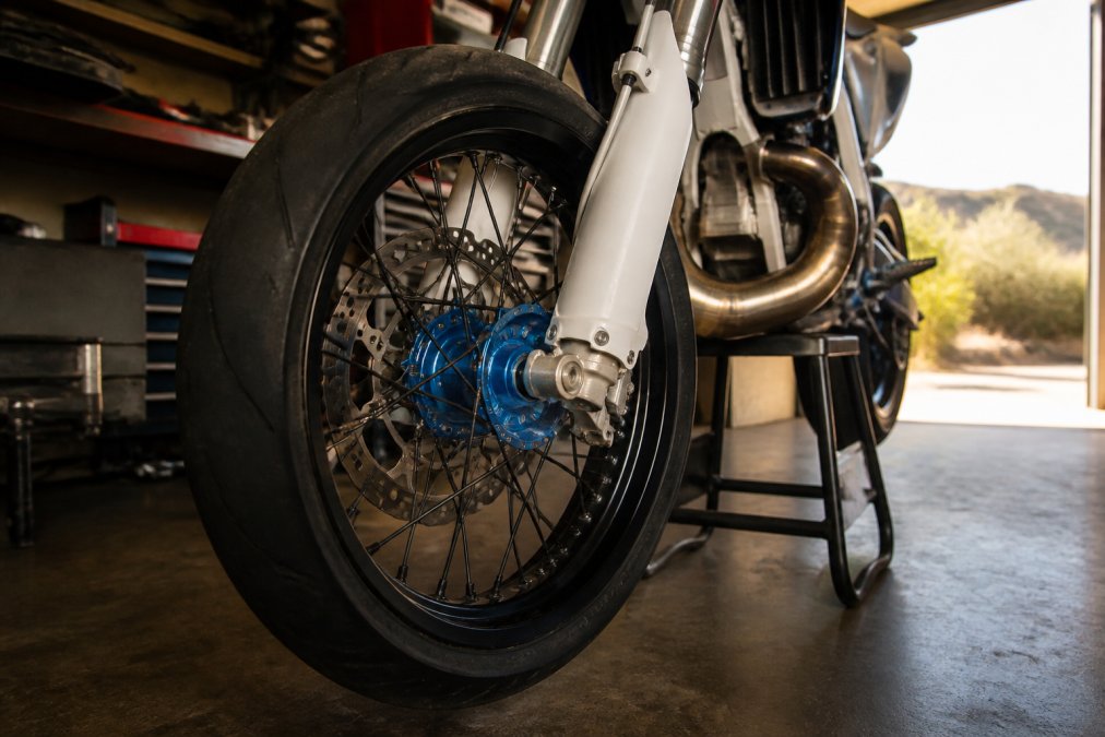 Close-up of a spoked supermoto wheel with a billet aluminum hub mounted on a dirt bike in a garage, morning light through an open door