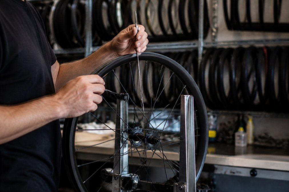 Close-up of a mechanic's hands tensioning a spoke on a mountain bike wheel in a workshop, with a truing stand and various carbon rims blurred in the background