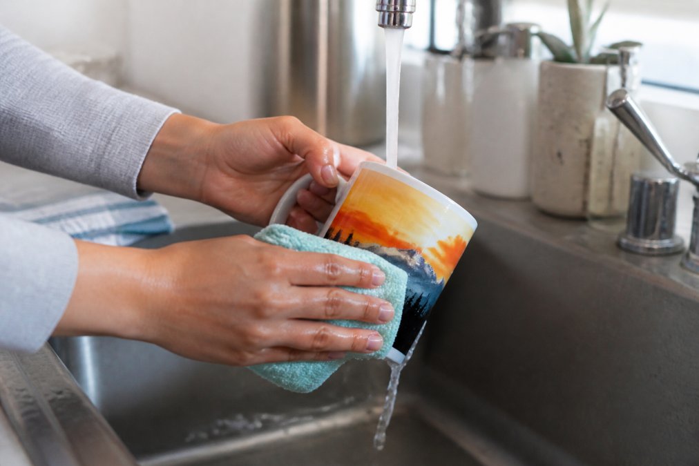 A custom ceramic mug being carefully hand-washed under warm running water with a soft cloth, showing proper gentle cleaning technique to preserve the print