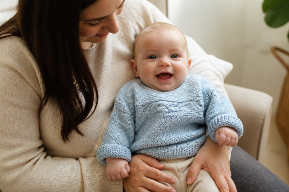 Parent holding a finished personalized hand-knitted baby sweater with their child's name embroidered across the front, showing the sweater on a real baby in soft natural lighting