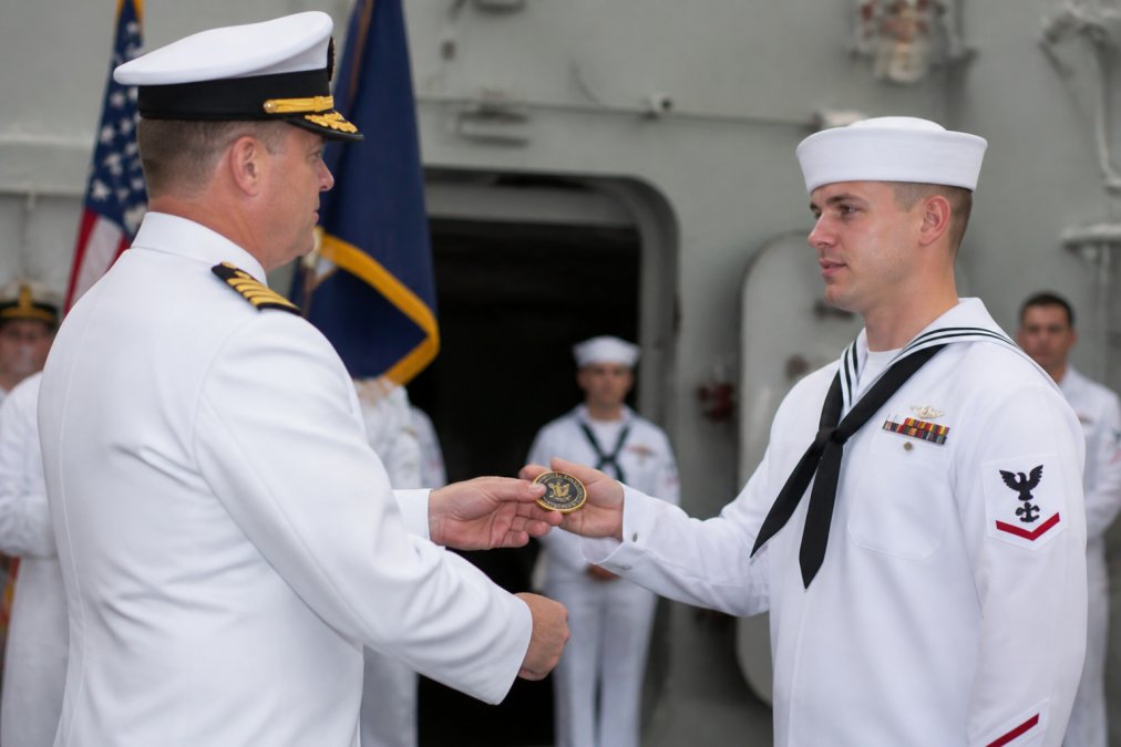 Naval officers in dress uniforms exchanging challenge coins during formal military ceremony, with commanding officer presenting coin to sailor, formal military setting with flags and formal lighting