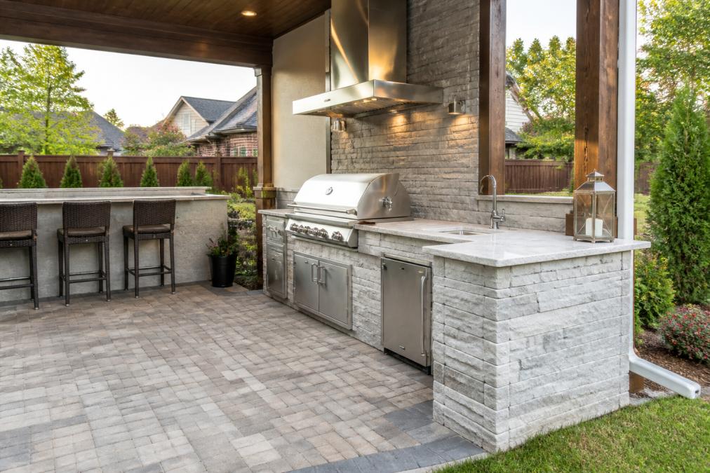 Professional outdoor kitchen installation showing custom stone masonry countertops, built-in stainless steel grill with proper ventilation, and integrated hardscape patio with permeable pavers and drainage grading