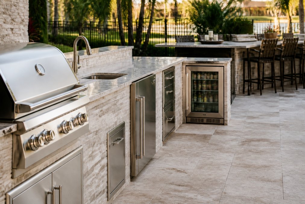 Close-up of built-in outdoor kitchen components including gas grill, stainless steel refrigerator, sink with faucet, and beverage center with bar seating in the background