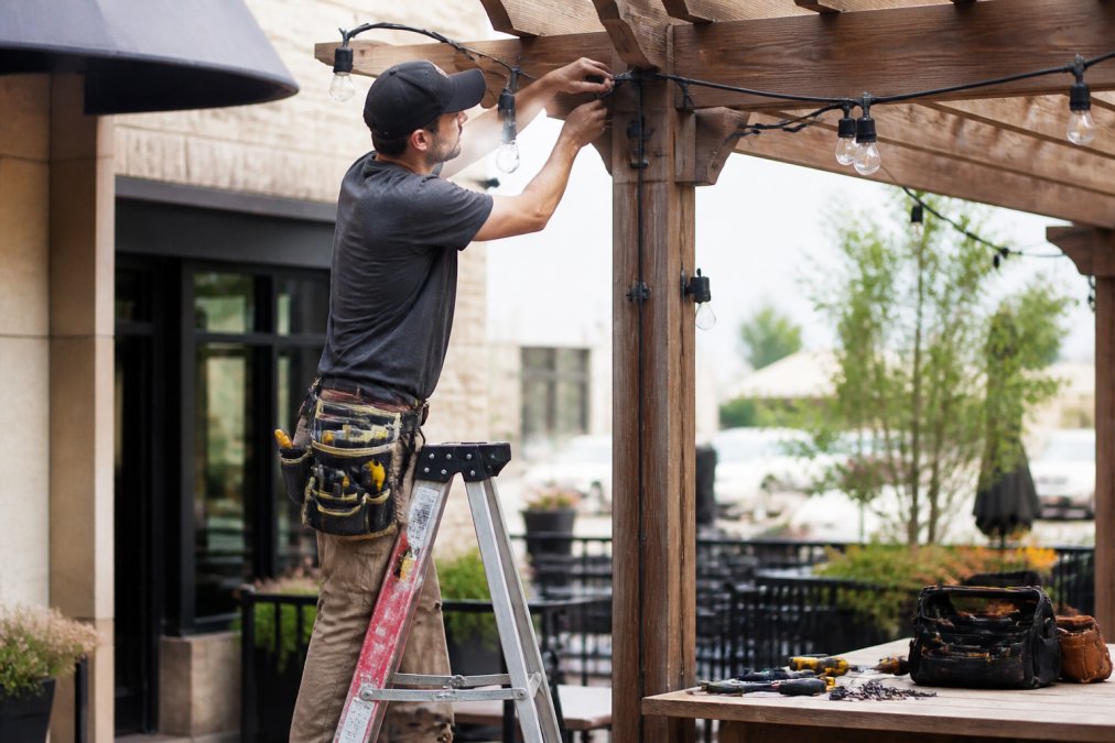A professional outdoor lighting installer on a ladder securing commercial-grade bistro string lights to a wooden pergola post on a small restaurant patio, with weatherproof connectors visible and tools on a nearby workbench