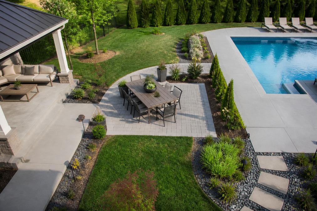 Overhead view of a completed backyard with distinct zones: a concrete patio with lounge seating on one side, a separate dining area with a table and chairs in the center, and a pool area with clear visual separation between spaces