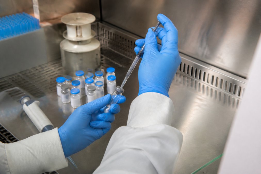 A gloved researcher in a laboratory hood carefully injecting bacteriostatic water into a small glass vial with a syringe, surrounded by labeled peptide vials and a pipette on a clean stainless steel bench