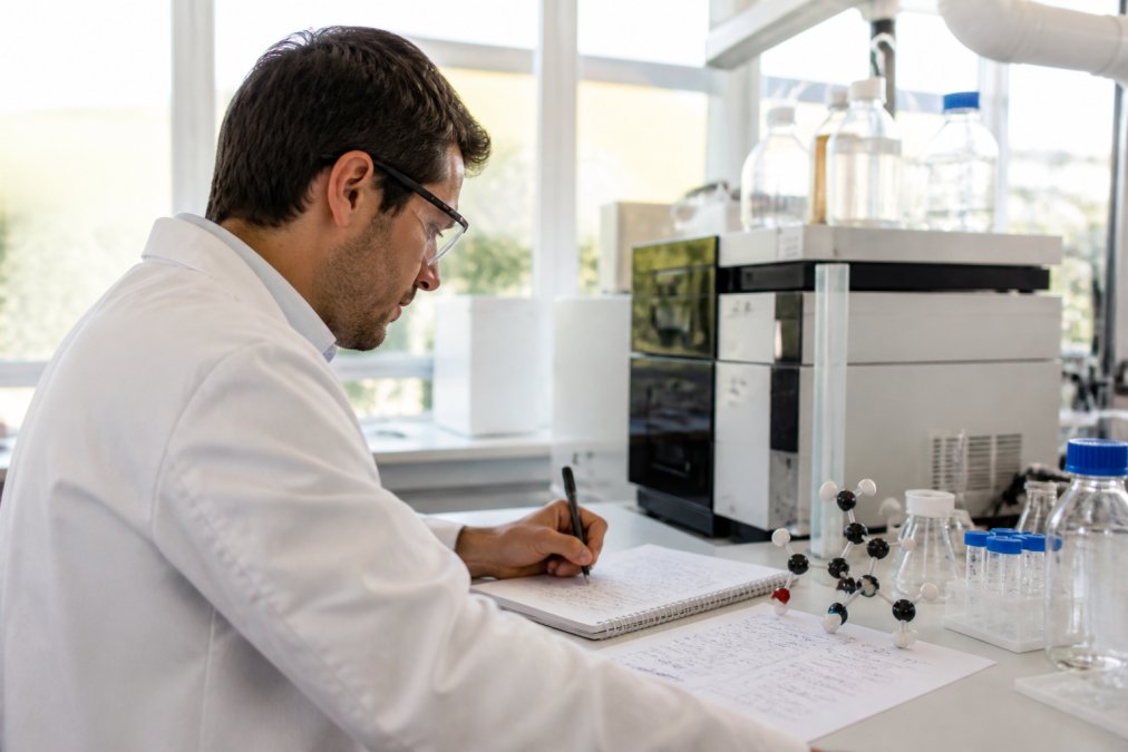 Researcher sitting at laboratory desk with laptop displaying amino acid formulas and molecular structures, with handwritten notes visible on desk, bright natural lab lighting