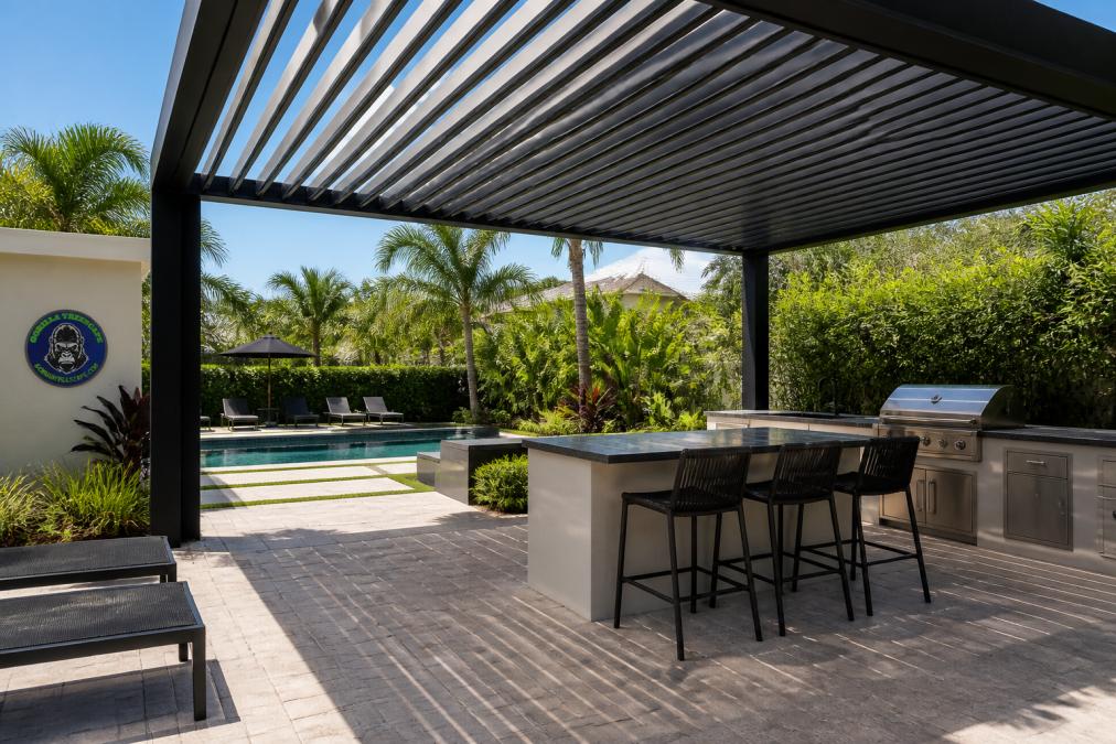 A modern aluminum louvered pergola installed over a paver patio in a South Florida backyard, with an outdoor kitchen and tropical landscaping visible in the background