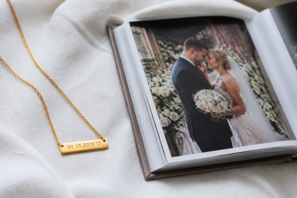 Close-up of an engraved gold necklace with a date inscription resting beside a printed custom photo book open to a wedding anniversary page, on a soft white linen surface
