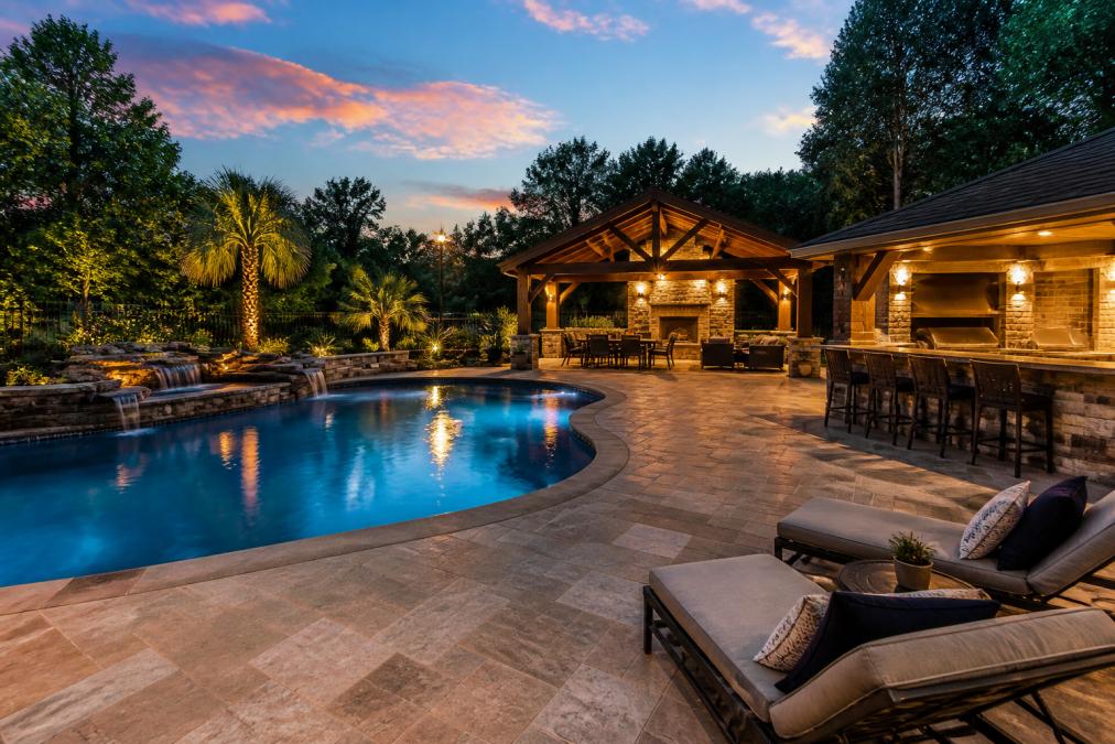 A wide shot of a newly finished backyard at dusk with a custom inground pool, stone patio, and outdoor kitchen area, warm lighting reflecting on the water