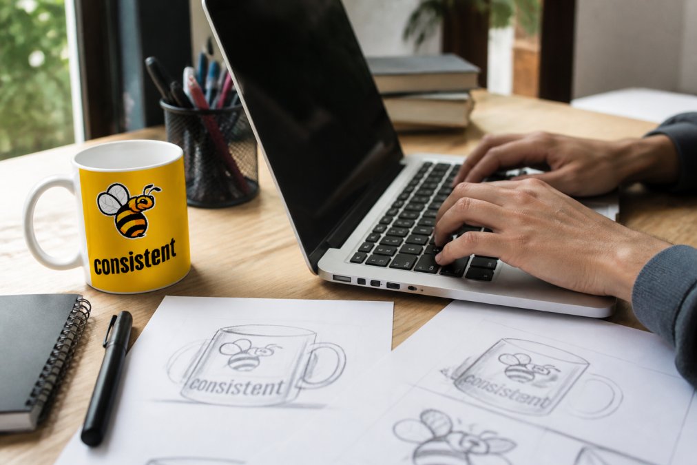 Hands using a laptop with design tool interface to customize a mug, coffee cup and design sketches visible on wooden desk with natural window lighting
