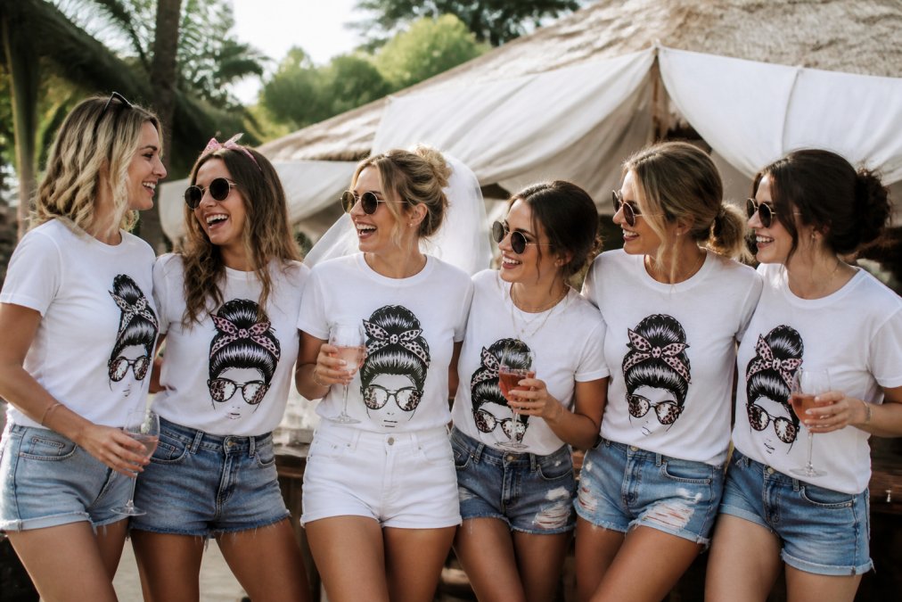 A group of women wearing matching custom graphic tees at a bachelorette party, laughing and holding drinks in a bright outdoor setting
