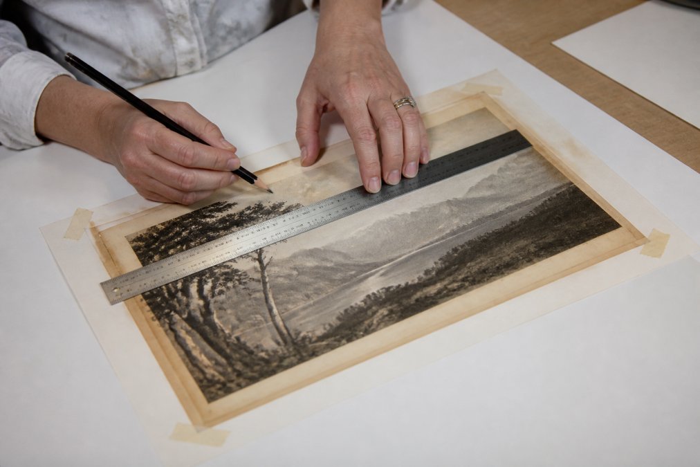Close-up of hands carefully measuring an antique print with a metal ruler and pencil on a clean, flat surface, showing the masking tape method securing corners of the artwork