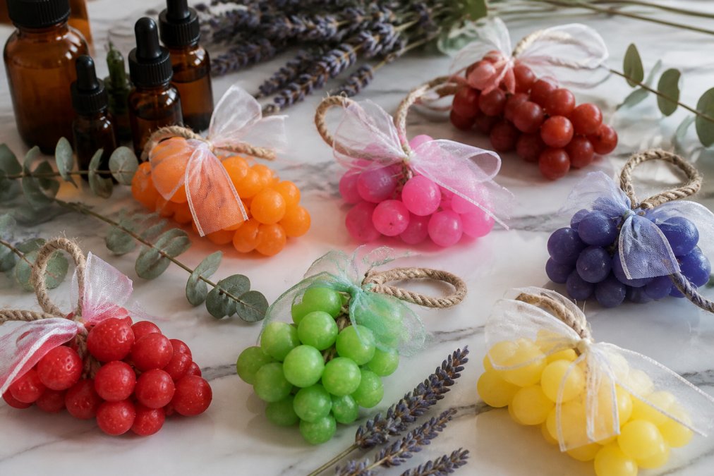 Close-up of artisanal soap bars in various natural colors, deep blues, warm terracottas, soft greens, arranged on a marble surface with dried lavender sprigs, eucalyptus leaves, and amber glass bottles of essential oils nearby