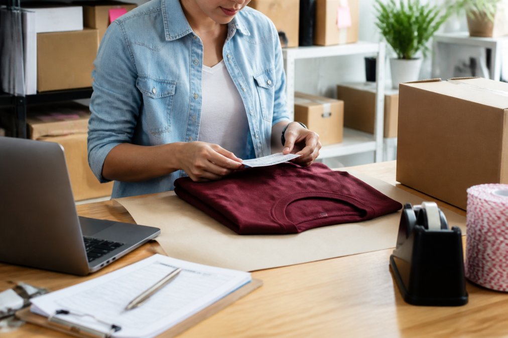 Person at desk with laptop and shipping label, preparing to package a custom t-shirt for return or exchange, with organized workspace showing order documentation