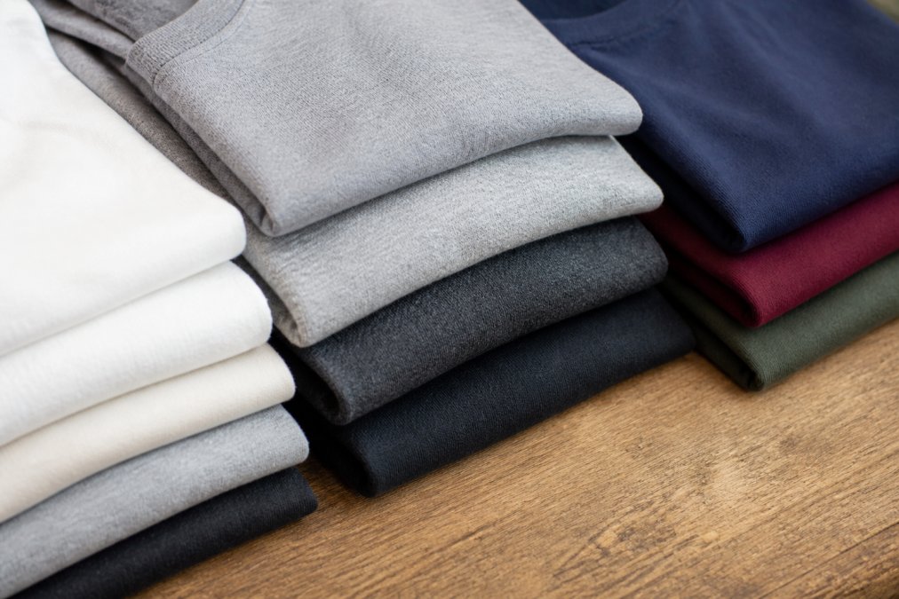 Close-up of several neatly folded t-shirts in different fabric weights and cotton blends arranged on a flat wooden surface, showing visible texture differences in natural daylight