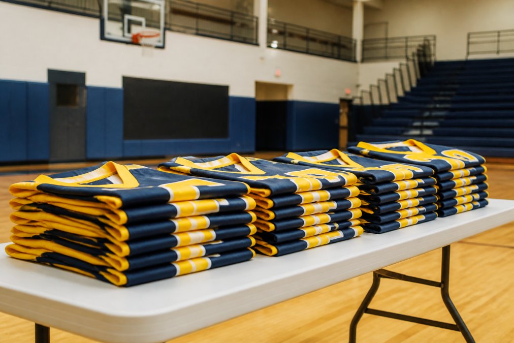 Neatly stacked custom team jerseys in matching colors arranged on a folding table in a gymnasium, bright overhead lighting, ready for distribution before a game