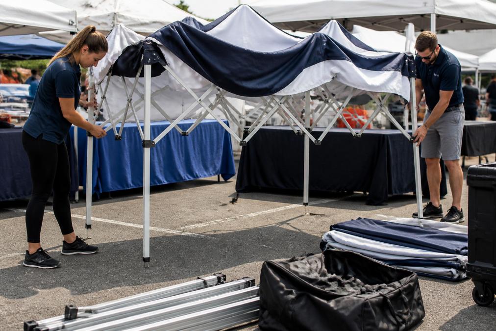 Event staff assembling a branded 10x10 pop-up canopy tent outdoors at a trade show, extending telescopic legs and positioning the dye-sublimated canopy top with company graphics visible