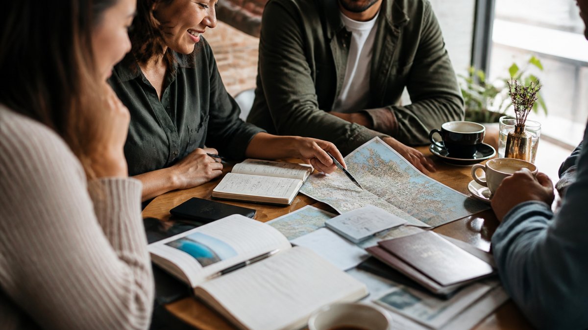 A diverse group of travelers reviewing a map and itinerary together at a café table, with notebooks and travel documents spread out, smiling and planning their trip