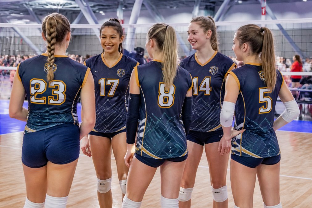 Professional youth volleyball team wearing custom sublimated jerseys in coordinated navy and gold colors, standing together on a court during a tournament, showing full designs with custom numbers and logos visible on front and back