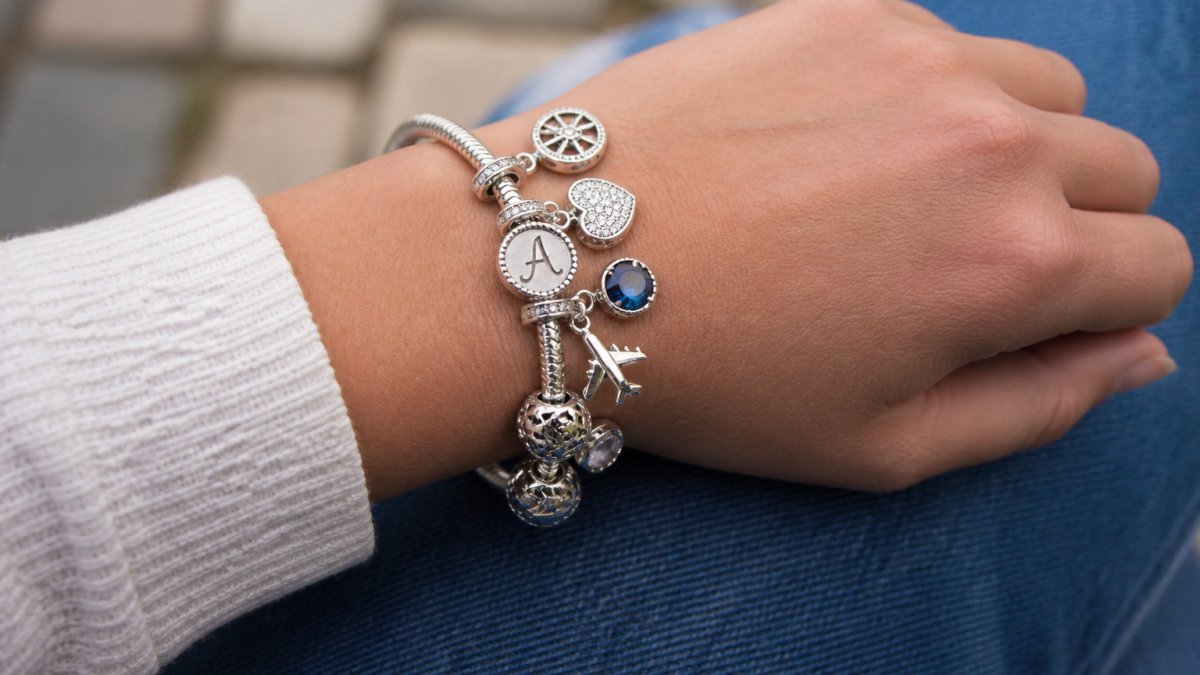 Close-up of a wrist wearing a custom sterling silver charm bracelet with varied charms including initials, birthstones, and travel-themed pendants in natural daylight