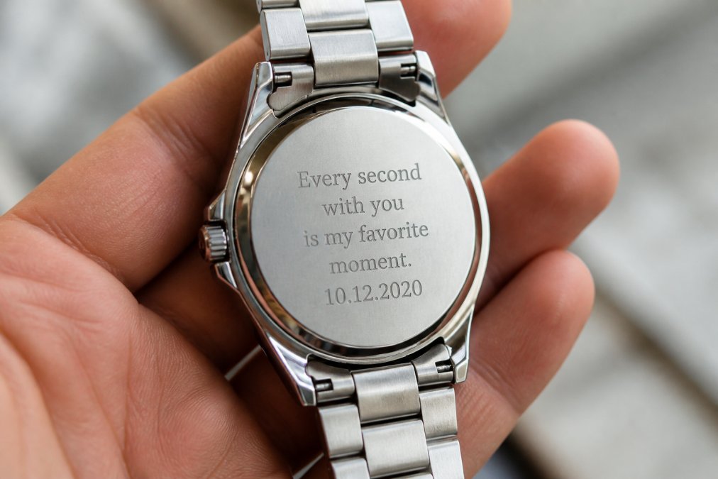 Macro shot of the back case of a stainless steel watch showing a freshly laser-etched personal message, held in a person's hand against a soft blurred background