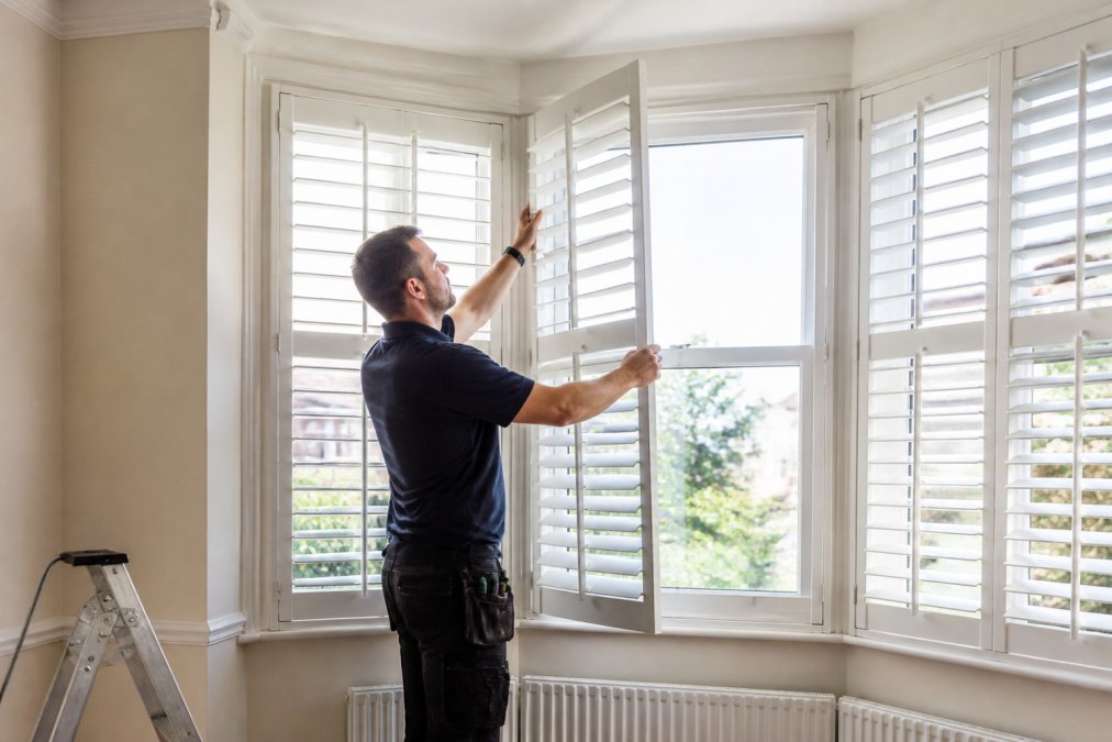 Professional craftsman installing custom white plantation shutters precisely into a period bay window frame, showing detailed measurement and fitting work