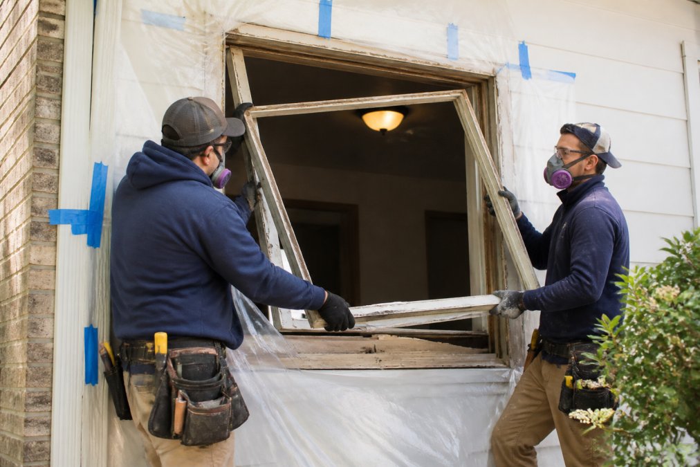 Professional installation crew carefully removing an old window frame from residential opening, with protective sheeting visible around work area and proper safety equipment in use