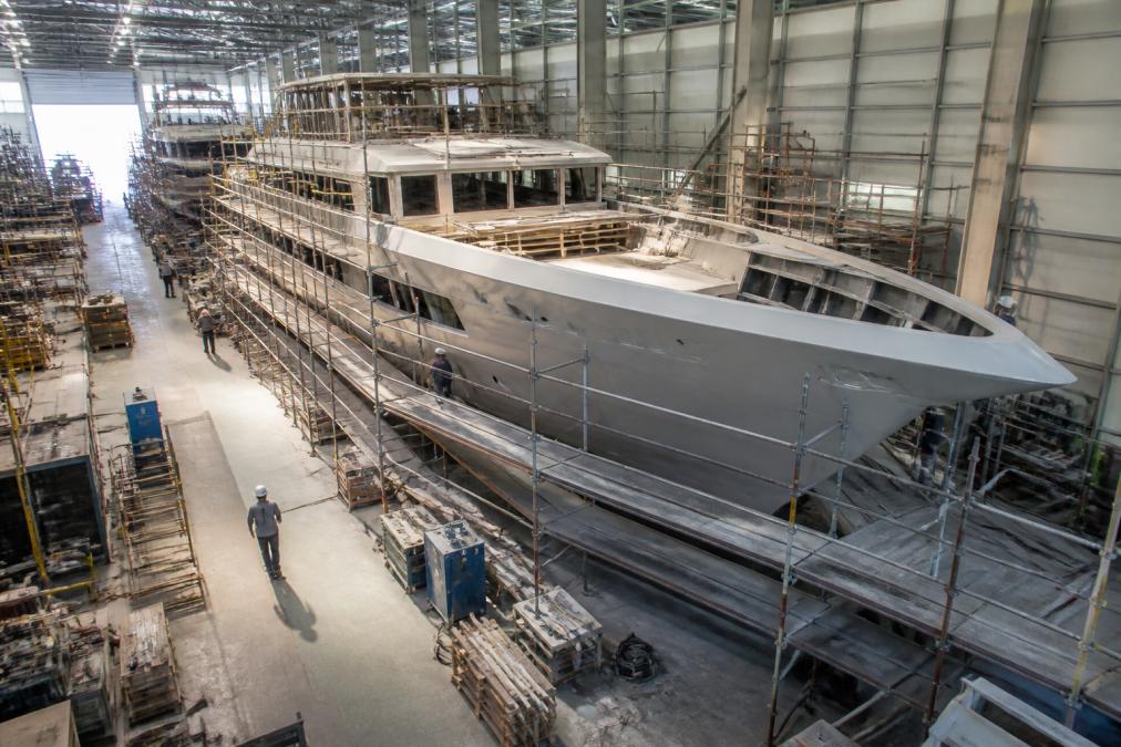 Modern shipyard facility with a large yacht hull under construction, showing welders working on steel sections, industrial cranes, and the vessel's curved hull taking shape in natural daylight
