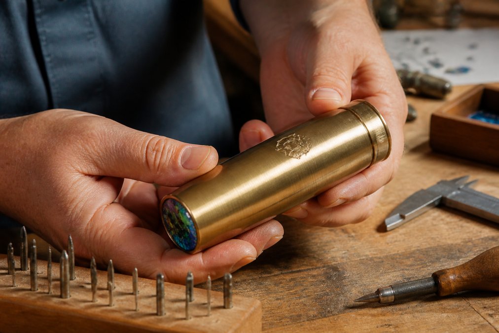 Close-up of hands holding a brass kaleidoscope with a custom logo printed on the exterior barrel, warm workshop lighting, wooden table with engraving tools nearby