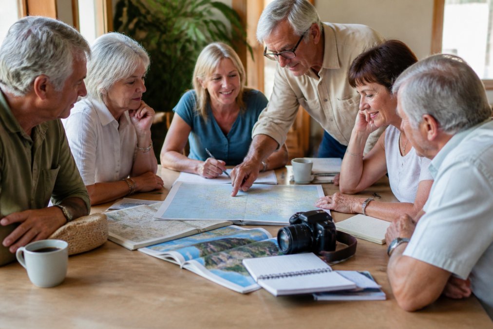A diverse group of mature travellers gathered around a table reviewing maps and itinerary notes, with a guide pointing to destinations, showing collaborative planning in action