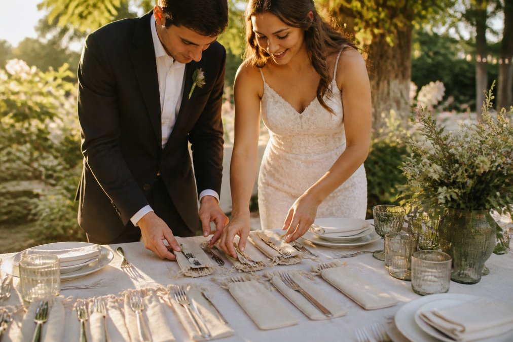 Bride and groom arranging reusable stainless steel cutlery sets with custom hemp pouches on an outdoor garden table, soft golden hour sunlight illuminating the place settings