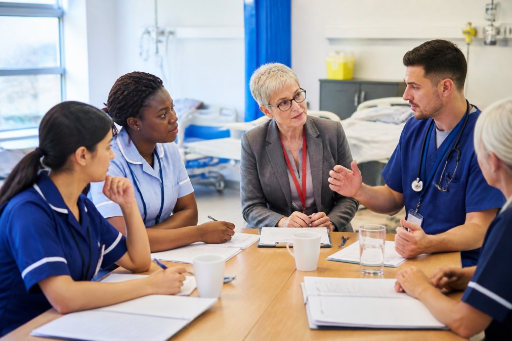 Healthcare professionals in modern training room collaborating around a table, discussing real-world clinical scenarios, diverse team with mix of nursing and administrative staff, natural light, engaged body language