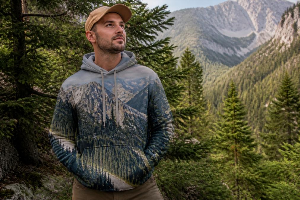 Person wearing a customized mountain view hoodie outdoors in natural lighting, standing near evergreen trees with the nature design clearly visible on the chest and shoulders