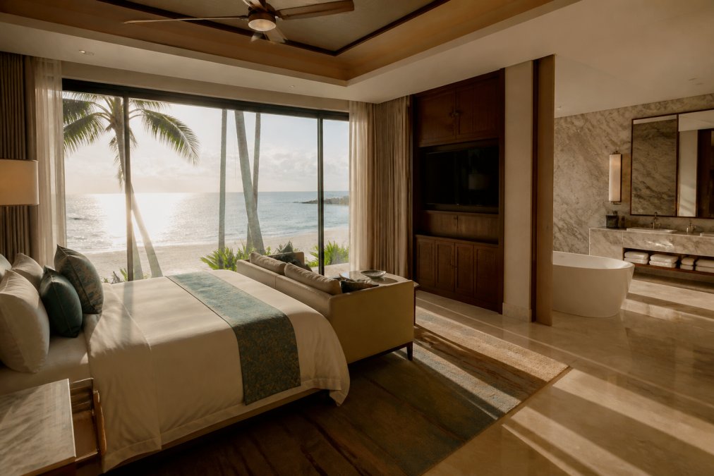 Luxury hotel suite with floor-to-ceiling windows overlooking a tropical beachfront, high-end furnishings, marble bathroom visible in background, soft afternoon sunlight streaming through windows
