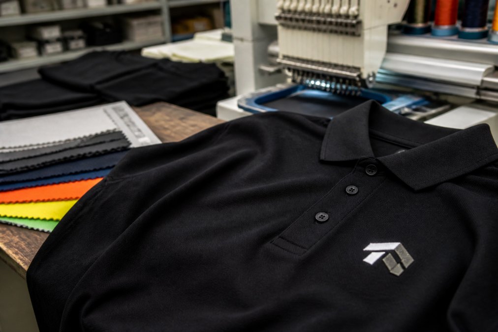Embroidered black polo shirt on a workbench with fabric swatches and an embroidery machine in a workshop