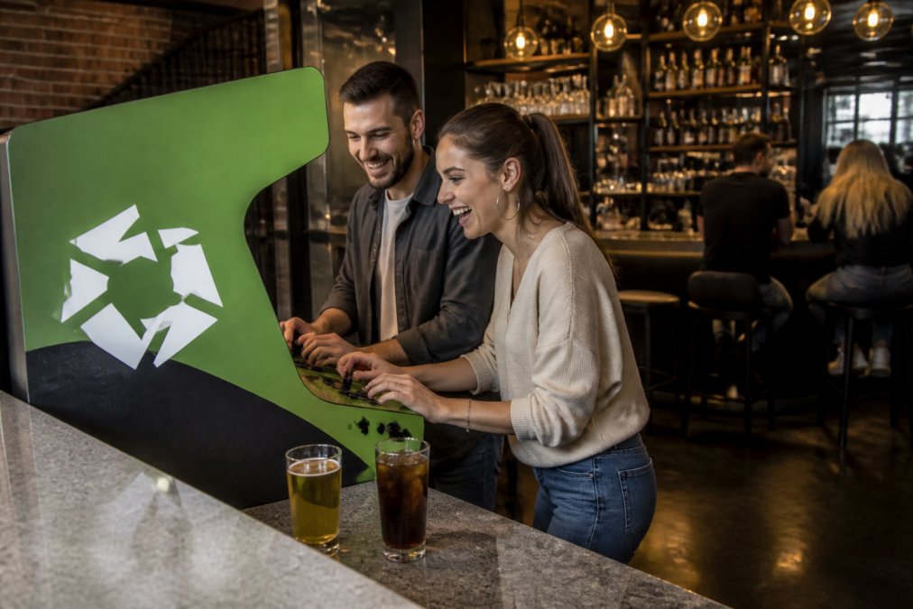A modern restaurant bar interior where a custom arcade cabinet displays vibrant brand colors and a glowing logo, with two customers laughing while playing and drinks on the counter nearby